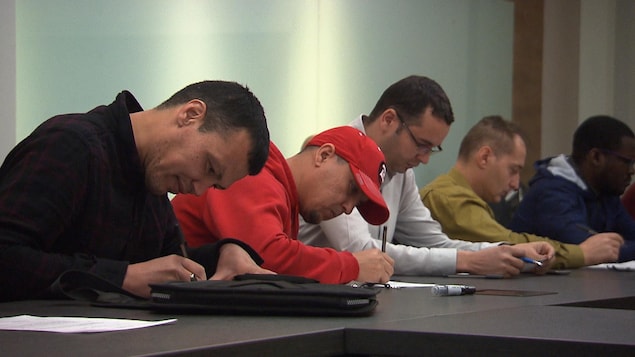 Des immigrants, tous des hommes, écrivent assis à une longue table.