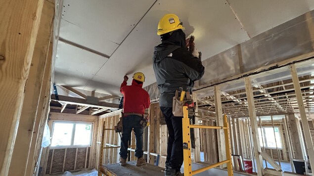 Deux hommes vissent des feuilles de gypse au plafond d'un édifice en construction.  Ils sont sur une plateforme surélevée.
 