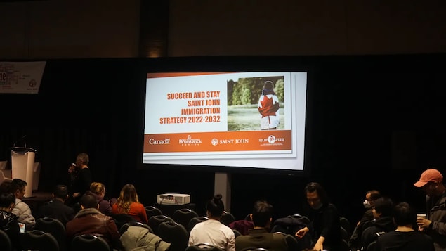 Mga tao na nakaupo at nakatingin sa projector kung saan naka-flash ang isang presentation na pinamagatang 'Succeed and Stay Saint John Immigration Strategy 2022-2023.'