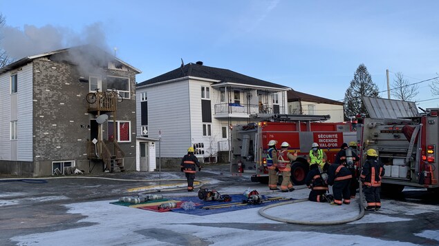De la fumée s'échappe du bâtiment. Huit pompiers sont près de leur camion dans la rue.