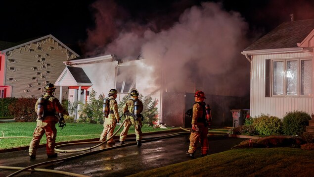Quatre pompiers devant les trois maisons sinistrées. L'un d'eux arrose la maison du centre avec un boyau.