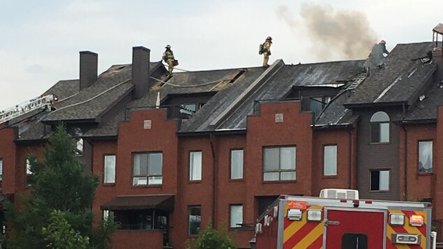 Un édifice à condos en flammes. Un véhicule de police et un camion de pompier sont devant l'édifice. Des pompiers sont sur le toit. Un édifice à condos en flammes. Un véhicule de police et un camion de pompier sont devant l'édifice. Des pompiers sont sur le toit.