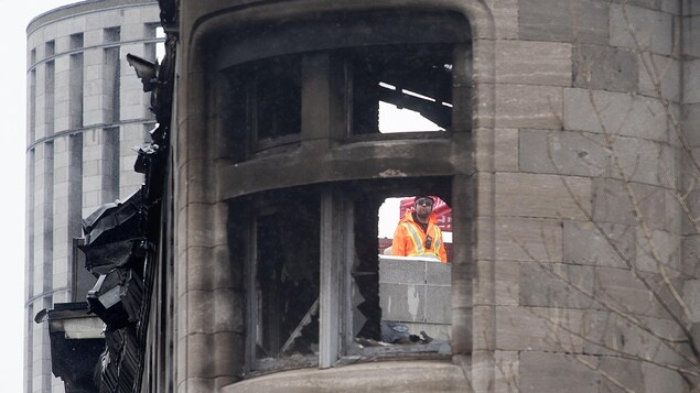 Un secouriste portant des verres fumés regarde à travers le trou béant formé par l'éclatement de la fenêtre d'une tourelle de l'immeuble patrimonial incendié.