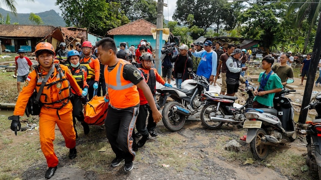 Dans un village où certaines maisons sont détruites, des secouristes transportent un corps dans un sac, entourés de badauds.