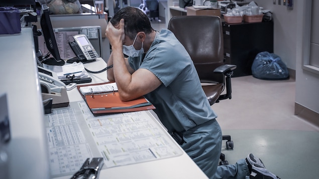Un médecin en milieu hospitalier à genoux par terre et accoudé à une table. Une main sur son front et l'autre tient le combiné d'un téléphone.