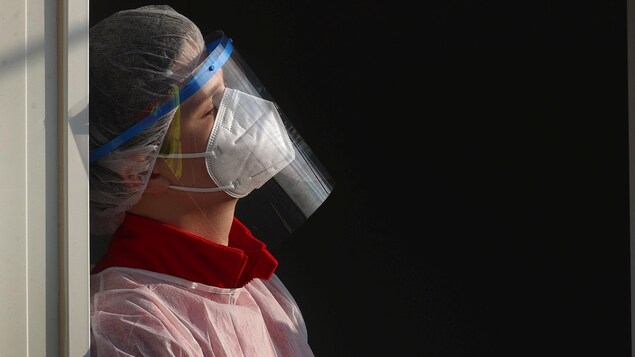 Une infirmière vêtue de l'équipement de protection personnelle est adossée contre un cadre de porte. Elle paraît exténuée et ferme les yeux.