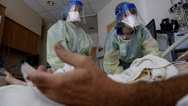 Deux infirmières inclinées devant un homme, dont on ne voit que le bras gauche, couché sur un lit d'hôpital.