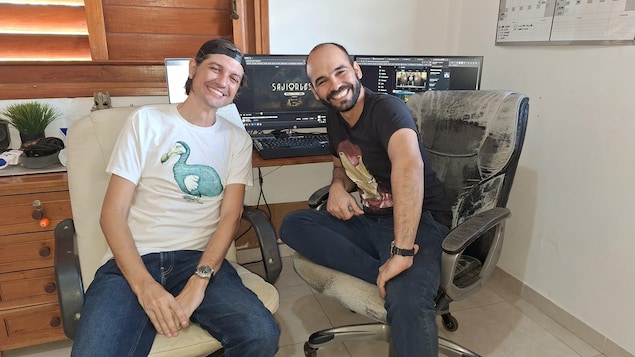 Josuhe Pagliery et David Darias, souriants, joignent le poing dans un moment de complicité, devant des écrans d'ordinateurs. 
