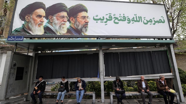 Des Iraniens sont assis à un arrêt de bus sous un panneau d'affichage représentant le défunt guide suprême iranien, l'ayatollah Rouhollah Khomeini (à gauche), son successeur, le défunt ayatollah Ali Khamenei (au centre), et le nouveau guide suprême Mojtaba Khamenei, à Téhéran, le 31 mars 2026.