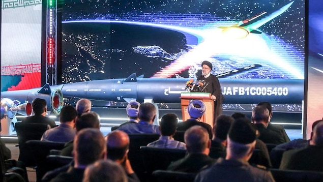 Un homme prononçant un discours devant un auditoire avec en arrière-plan un missile.