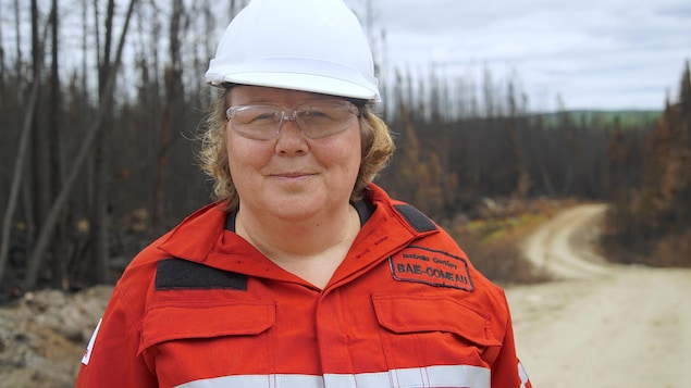 On voit une femme qui porte un casque de sécurité et un vêtement de sécurité. Elle se trouve sur les lieux d'un incendie de forêt. Au loin, on aperçoit les arbres calcinés d'une forêt. On voit une femme qui porte un casque de sécurité et un vêtement de sécurité. Elle se trouve sur les lieux d'un incendie de forêt. Au loin, on aperçoit les arbres calcinés d'une forêt.