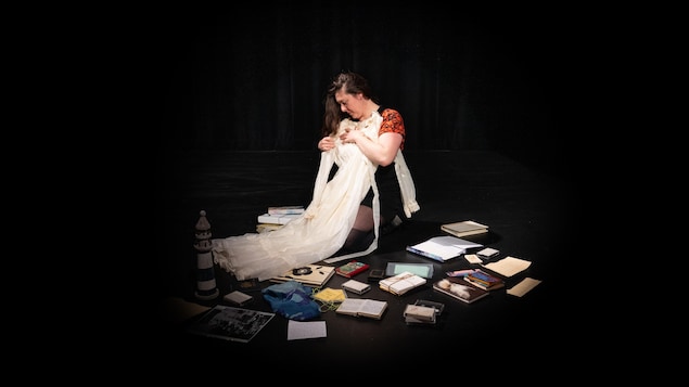 Sur une scène, Isabelle Rivest, assise, tient une robe de mariée, entourée de cassettes, photos et livres étalés par terre.