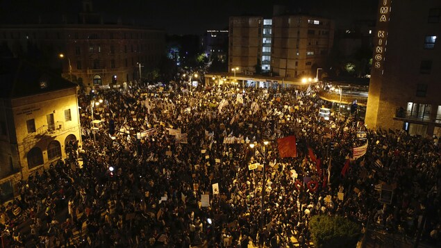 Une foule dense de milliers de manifestants dans les rues le soir, portant des pancartes et des drapeaux.