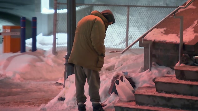 Une personne en situation d’itinérance trimballant des effets personnels dans des sacs de plastique se tient devant le refuge Lauberivière un soir d’hiver. 