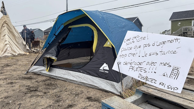 Une tente de camping est installée sur la plage à côté d'une pancarte où il est écrit « C'est chez nous icitte! Parce que pas d'logement, trop cher! Je suis tanné de déménager avec mes palettes!!! Respectez moi svp.»