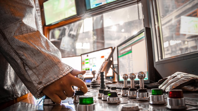La main d'un homme manipule une manette dans la salle de contrôle d'une aciérie où se trouve plusieurs boutons et écrans.