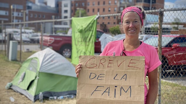 Jacinthe Comeau tient une affiche avec l'inscription «Grève de la faim», avec sa tente en arrière-plan.