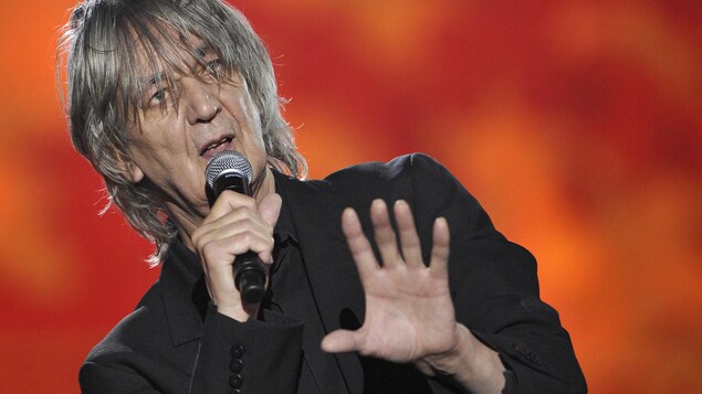 Le chanteur a les cheveux dans les yeux et tient un micro. Le chanteur a les cheveux dans les yeux et tient un micro.