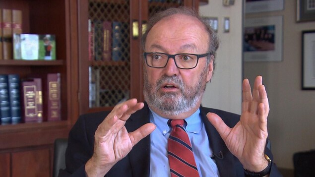 Jacques R. Fournier gesticule devant une bibliothèque remplie de livres. Jacques R. Fournier gesticule devant une bibliothèque remplie de livres.