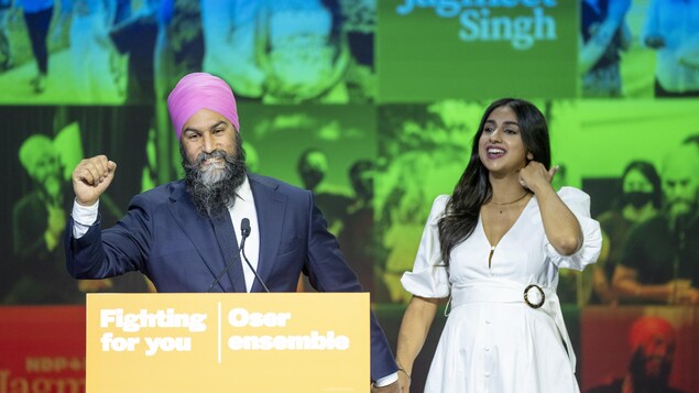 Jagmeet Singh lève le poing en souriant. Il tient la main de son épouse devant un podium où est écrit le slogan « Fighting for you; Oser ensemble ».
