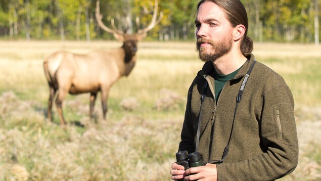 Jake Paleczny, des jumelles à la main, devant un caribou qui est en flou, en arrière plan. Jake Paleczny, des jumelles à la main, devant un caribou qui est en flou, en arrière plan.