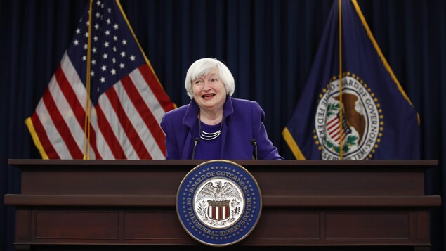 Janet Yellen est derrière un lutrin portant le sceau de la Fed, et devant des drapeaux des États-Unis et de la Fed.