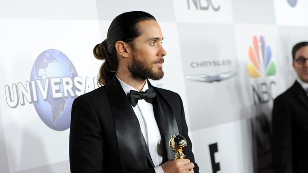 Un homme habillé en smoking pose pour une photo Oscar en main devant une bannière portant les logos des commanditaires d'un festival. Un homme habillé en smoking pose pour une photo Oscar en main devant une bannière portant les logos des commanditaires d'un festival.