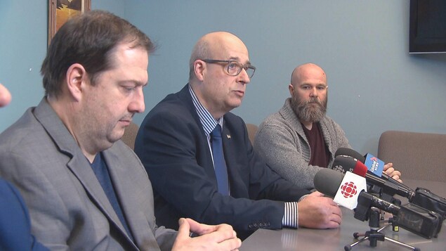 Trois hommes de profil parlent devant des micros posés sur une table.