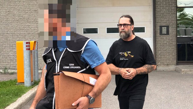Un homme barbu avec les cheveux longs attachés et des lunettes marche derrière un policier, menottes aux poignets Un homme barbu avec les cheveux longs attachés et des lunettes marche derrière un policier, menottes aux poignets