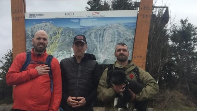 Trois hommes en haut d'une montagne devant une affiche du Mont-Saint-Anne.