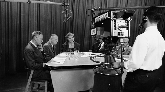 Dans un studio de télévision, réunis autour d'une table, Paul Sauriol, Roger Champoux, Jeanne Sauvé, Lucien Desbiens (caché derrière la caméra) et Réal Benoît. Dans le plan, un micro sur perche et un caméraman derrière sa caméra sur trépied. Dans un studio de télévision, réunis autour d'une table, Paul Sauriol, Roger Champoux, Jeanne Sauvé, Lucien Desbiens (caché derrière la caméra) et Réal Benoît. Dans le plan, un micro sur perche et un caméraman derrière sa caméra sur trépied.