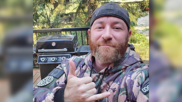 Jeremy MacKenzie, vêtu d'une combinaison de camouflage et coiffé d'une casquette à l'envers, prend un selfie à l'extérieur devant un barbecue et fait un signe de shaka avec son pouce et son auriculaire.