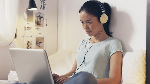 Une jeune fille est assise sur un lit, ordinateur portable sur les genoux et écouteurs sur les oreilles.