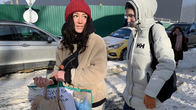 On voit une jeune qui tient des sacs, dans une rue enneigée. Elle tourne la tête de côté pour parler à son interlocuteur. Une autre jeune la suit de près.
