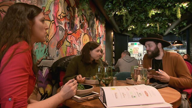 Trois personnes mangent un repas dans un restaurant, avec une grande peinture sur le mur derrière eux.