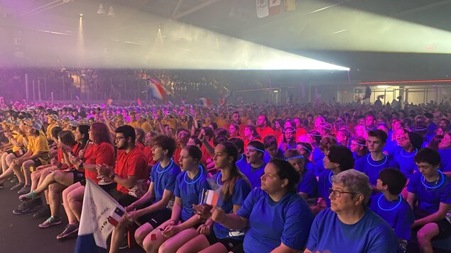 Des gens assis sur des chaises dans le centre sportif. On a regroupé ensemble les gens qui sont habillés en bleu, ceux qui sont habillés en rouge, ceux qui sont habillés en jaune, et ainsi de suite.