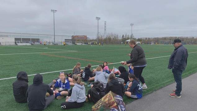 Des jeunes assis par terre sur un terrain de football synthétique extérieur pour manger des sandwiches.