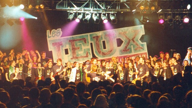 Une foule de jeunes participants sur scène devant une foule nombreuse dans la salle. Au fond de la scène, on peut lire Les Jeux. Une foule de jeunes participants sur scène devant une foule nombreuse dans la salle. Au fond de la scène, on peut lire Les Jeux.