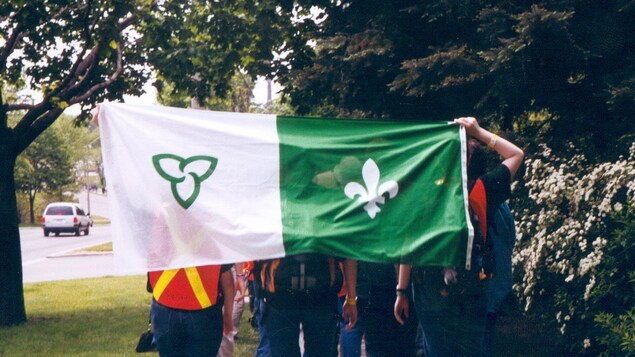 Le groupe est de dos à la caméra et marche sur un trottoir de la Ville Reine. Deux personnes tiennent un drapeau franco-ontarien dans leur dos. Le groupe est de dos à la caméra et marche sur un trottoir de la Ville Reine. Deux personnes tiennent un drapeau franco-ontarien dans leur dos.