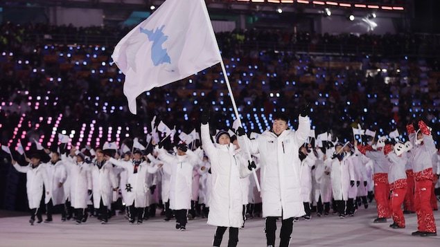 Les athlètes des deux Corées, vêtus de blanc, défilent derrière la drapeau de l'unification, une silhouette bleu pâle sur un fond blanc, portée par la Nord-Coréenne Hwang Chung-gum et le Sud-Coréen Won Yun-jong. Les athlètes des deux Corées, vêtus de blanc, défilent derrière la drapeau de l'unification, une silhouette bleu pâle sur un fond blanc, portée par la Nord-Coréenne Hwang Chung-gum et le Sud-Coréen Won Yun-jong.