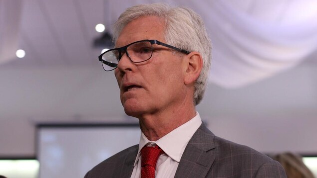 Jim Carr se tient debout, sérieux, la bouche légèrement entrouverte.