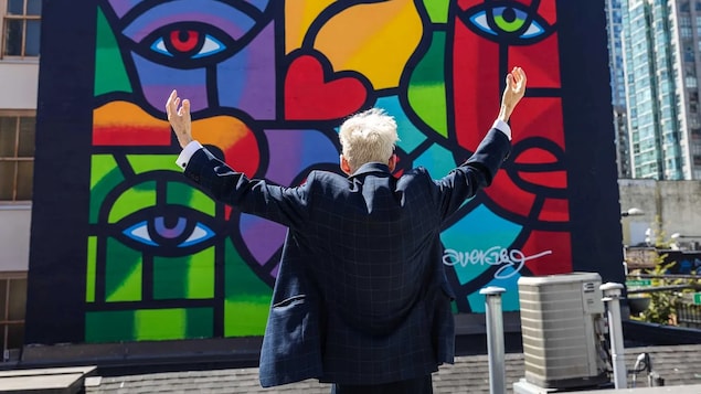 Joe Average, les bras grands ouverts devant sa murale « 40 years later », à Vancouver.