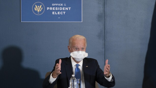 Joe Biden porte un énorme masque et est assis sous une pancarte disant : Bureau du président désigné.