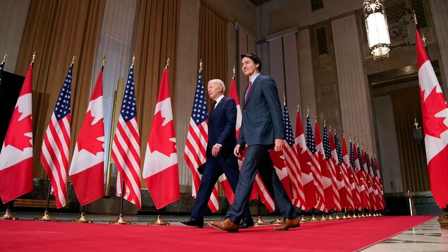 Dalawang lalaki na naglalakad sa red carpet sa tabi ng mga watawat ng Estados Unidos at Canada.