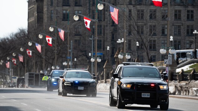 Des voitures de police dans une rue du centre-ville d'Ottawa où l'on voit des drapeaux canadiens et américains suspendus aux lampadaires.
