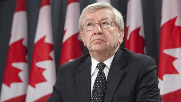 Un homme avec des lunettes, un veston et une cravate devant des drapeaux du Canada.