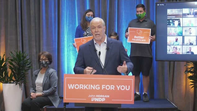 Derrière John Horgan, des sympathisants brandissent des pancartes demandant un meilleur service de santé, à côté d'un écran diffusant une conférence simultanée avec les candidats néo-démocrates.