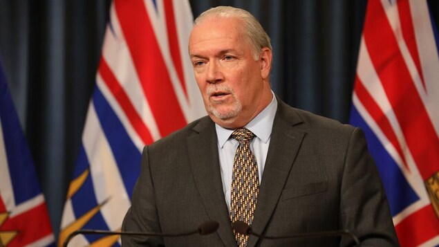Le premier ministre de la Colombie-Britannique, John Horgan pose de face devant deux micros. Il a un air sérieux.