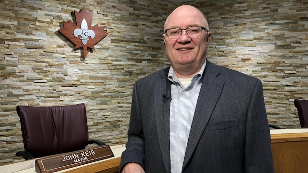 Un homme souriant en veston et portant des lunettes se tient devant un bureau et un logo d'une fleur de lys argent à l'intérieur d'une feuille d'érable rouge. Un homme souriant en veston et portant des lunettes se tient devant un bureau et un logo d'une fleur de lys argent à l'intérieur d'une feuille d'érable rouge.