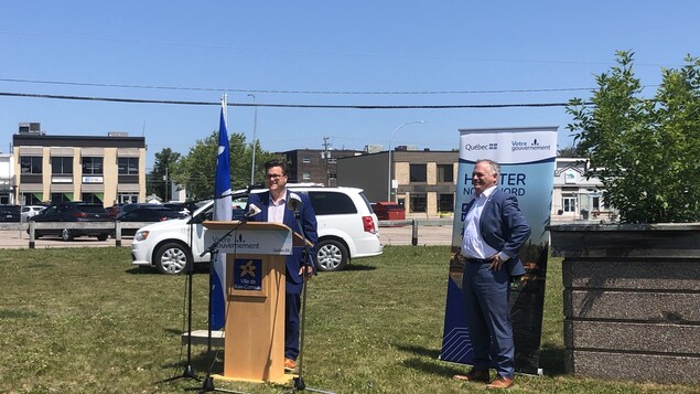 Yves Montigny et Jonathan Julien sont tous les deux devant un podium avec un drapeau du Québec derrière eux, ils se trouvent sur un espace vague vert.